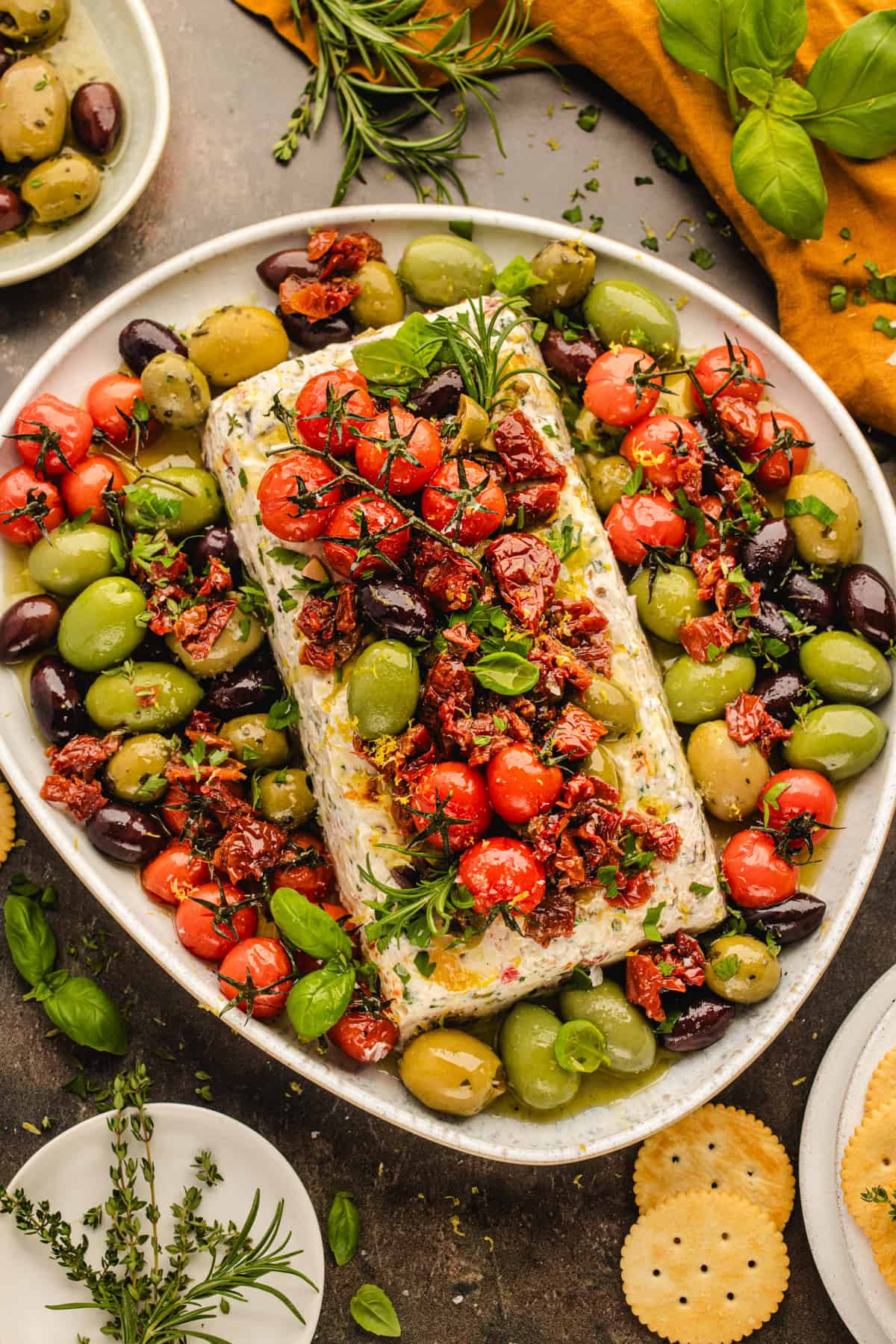 A platter with herbed cheese log, assorted green and black olives, cherry tomatoes, sun-dried tomatoes, and fresh herbs.