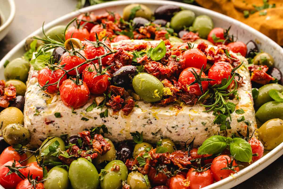 A rectangular block of herbed cheese topped with roasted cherry tomatoes, sundried tomatoes, olives, fresh herbs.