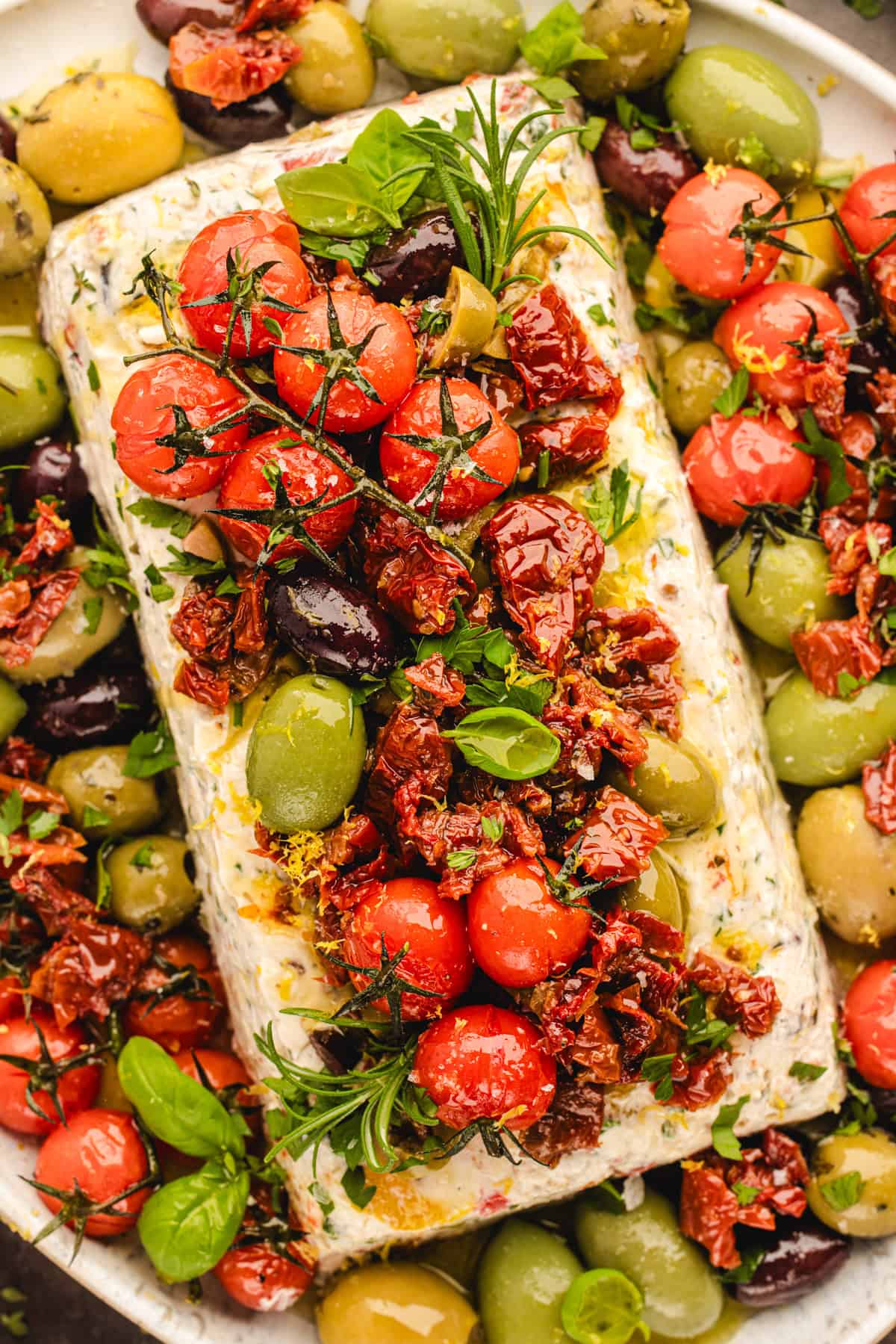 A rectangular block of herbed cheese topped with cherry tomatoes, sun-dried tomatoes, olives, and fresh herbs.