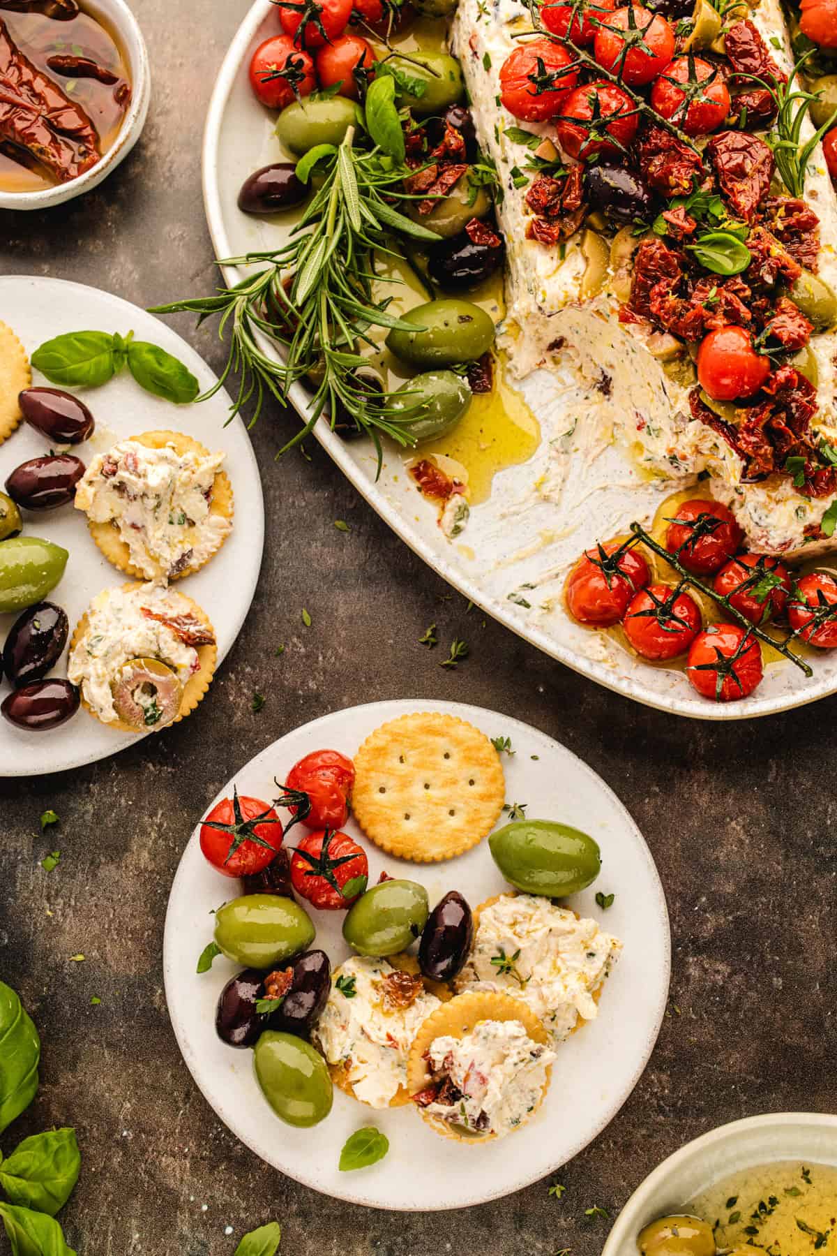 Plates with creamy cheese spread, crackers, olives, roasted tomatoes, and herbs. A large platter holds more cheese spread, tomatoes, olives, sun-dried tomatoes, and fresh rosemary. 