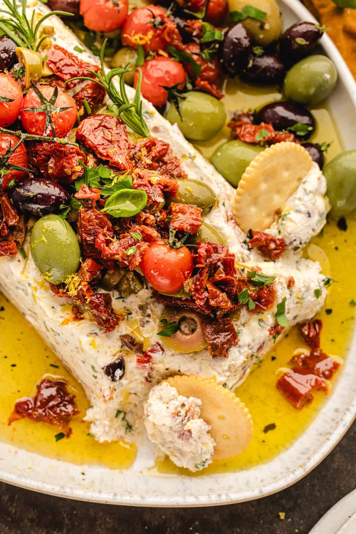 A creamy cheese log topped with olives, cherry tomatoes, sun-dried tomatoes, and herbs.