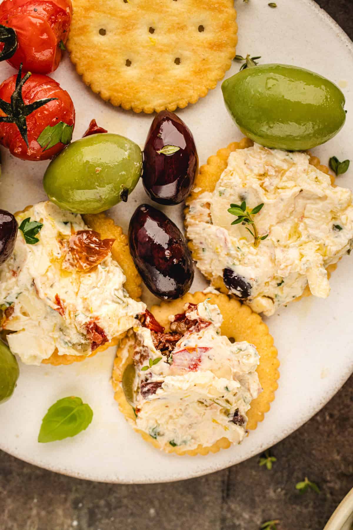 A plate with crackers topped with herbed cheese spread, black and green olives, roasted tomatoes, and fresh thyme for garnish.