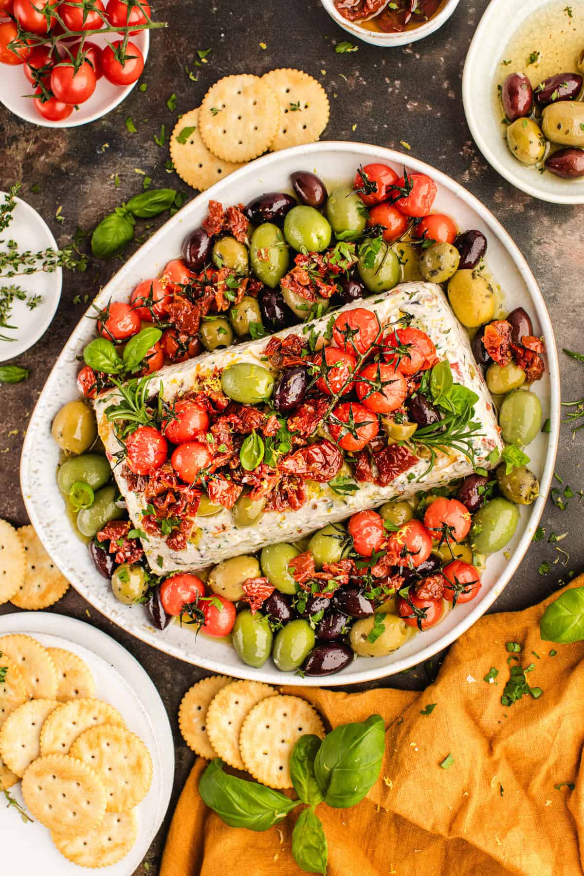A platter with herbed cheese log, assorted olives, cherry tomatoes, sun-dried tomatoes, fresh herbs, and round crackers.