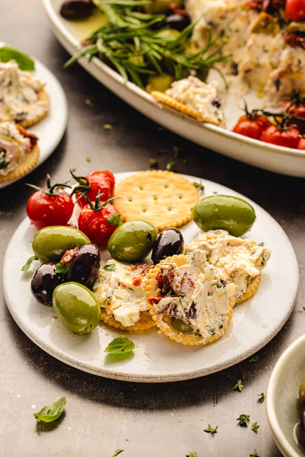 A small plate with crackers topped with herbed cheese, green and black olives, and roasted cherry tomatoes, garnished with fresh herbs.
