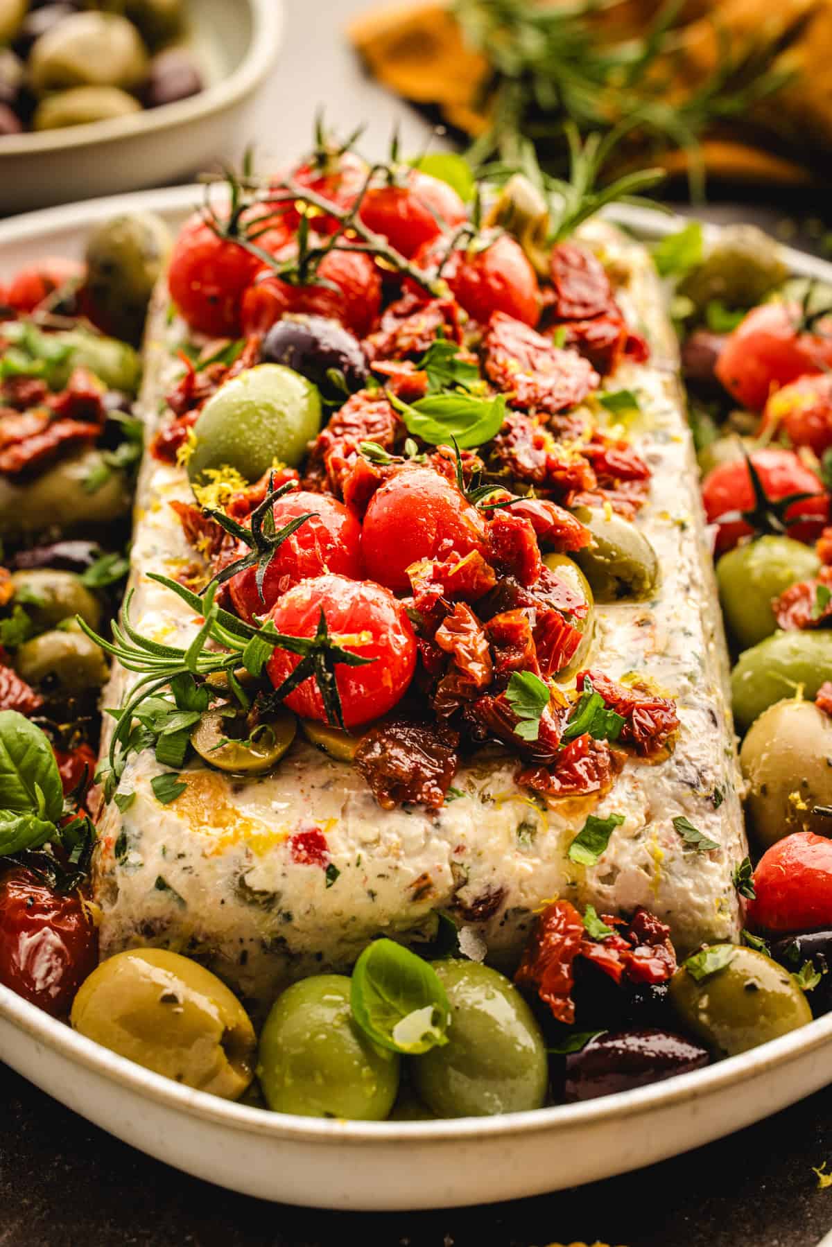 A rectangular cheese log topped with cherry tomatoes, sun-dried tomatoes, herbs.