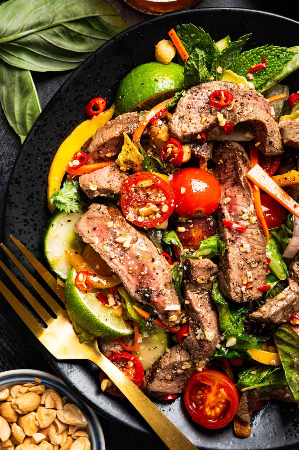 Thai Beef Salad A colorful Thai beef salad with grilled steak slices, cherry tomatoes, cucumbers, carrots, herbs, lime wedges, and red chili.