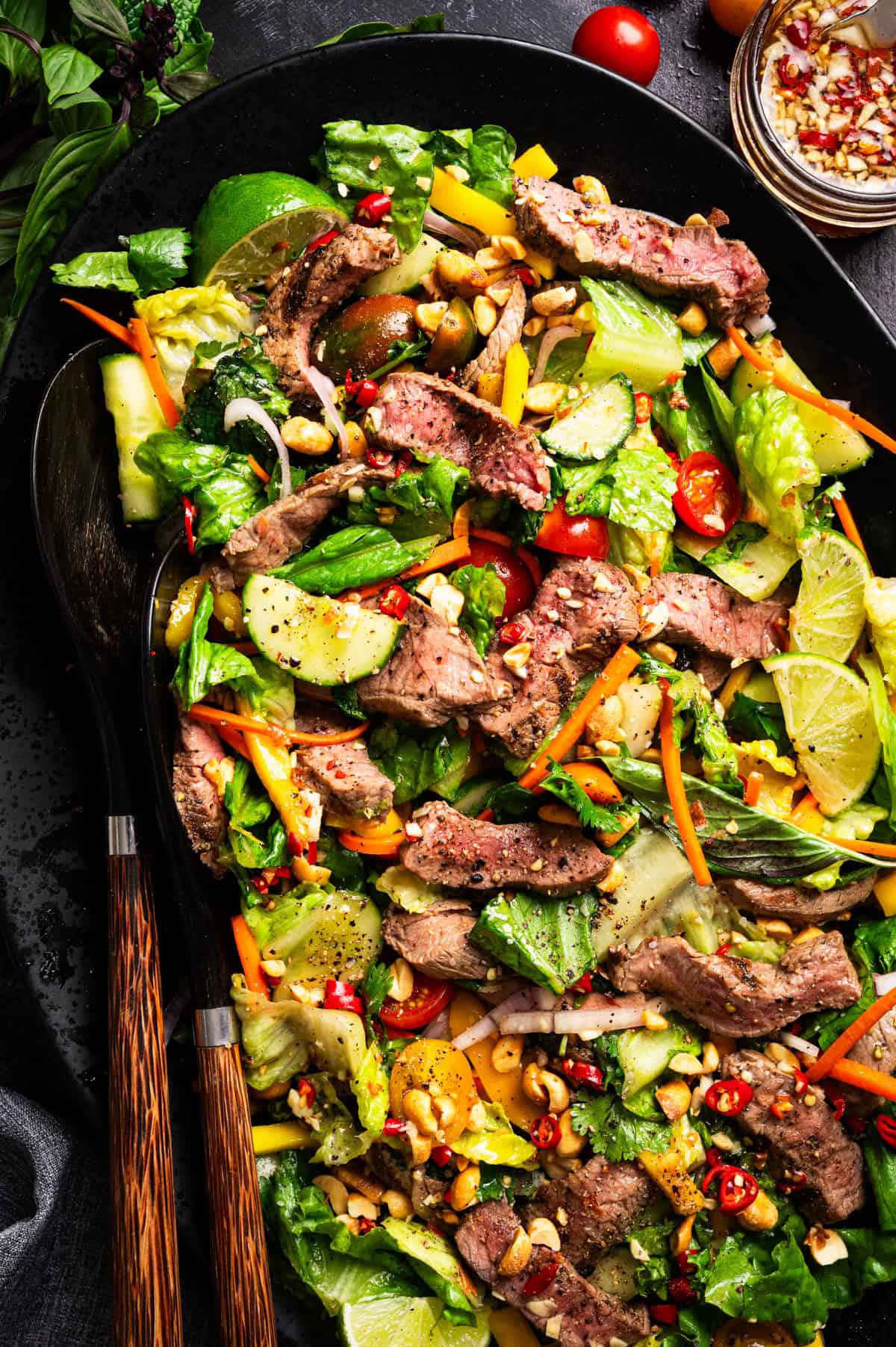 Thai Beef Salad Colorful Thai beef salad with grilled steak slices, mixed greens, carrots, cucumber, cherry tomatoes, chili, lime wedges, and fresh herbs, served in a black dish.