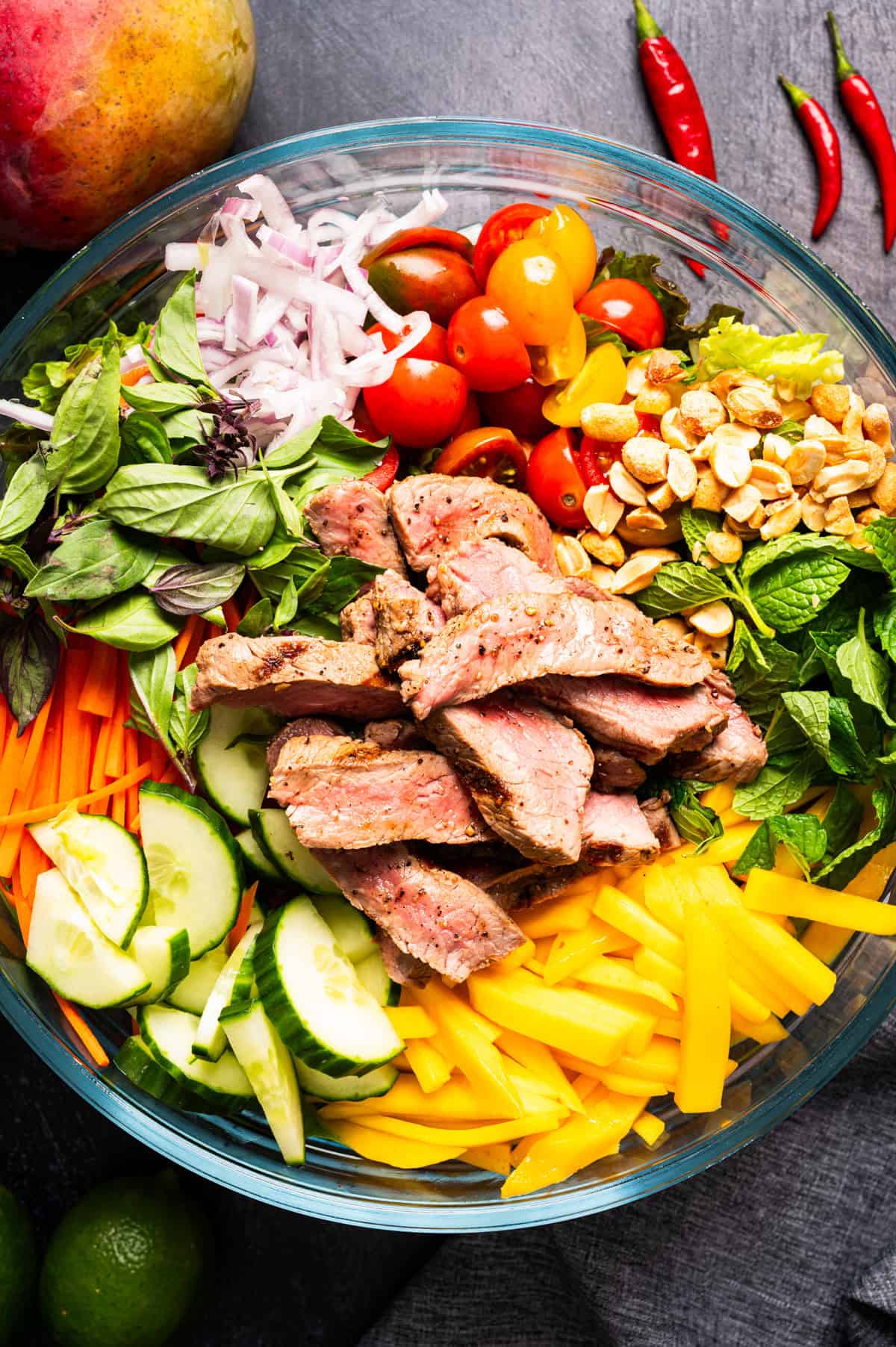 Thai Beef Salad A glass bowl filled with sliced steak, cucumbers, carrots, cherry tomatoes, mango, basil, mint, red onions, peanuts, and chili peppers, arranged in colorful sections.
