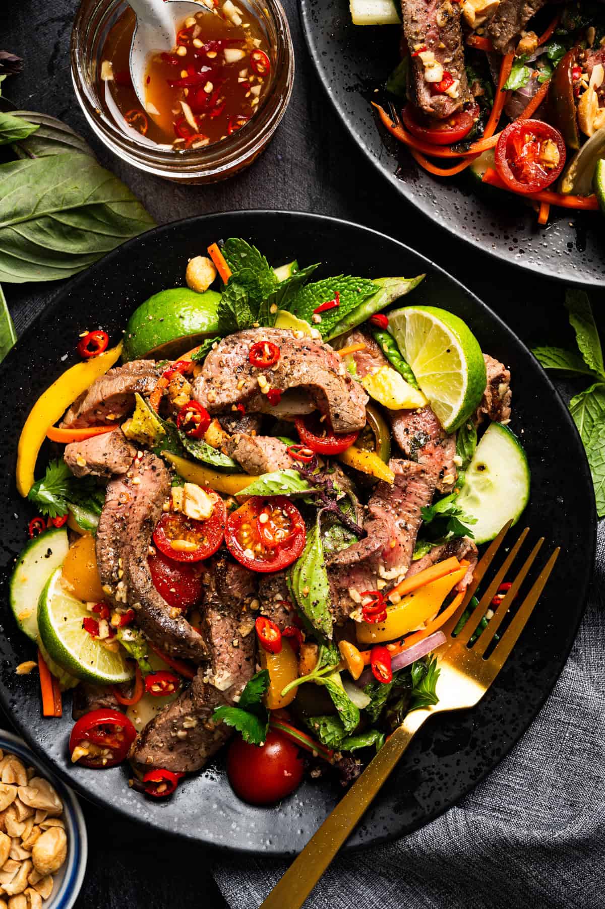 Thai Beef Salad A vibrant Thai beef salad with grilled steak slices, mixed greens, cucumbers, tomatoes, bell peppers, lime wedges, herbs, sliced chilies, and peanuts, served on a black plate.