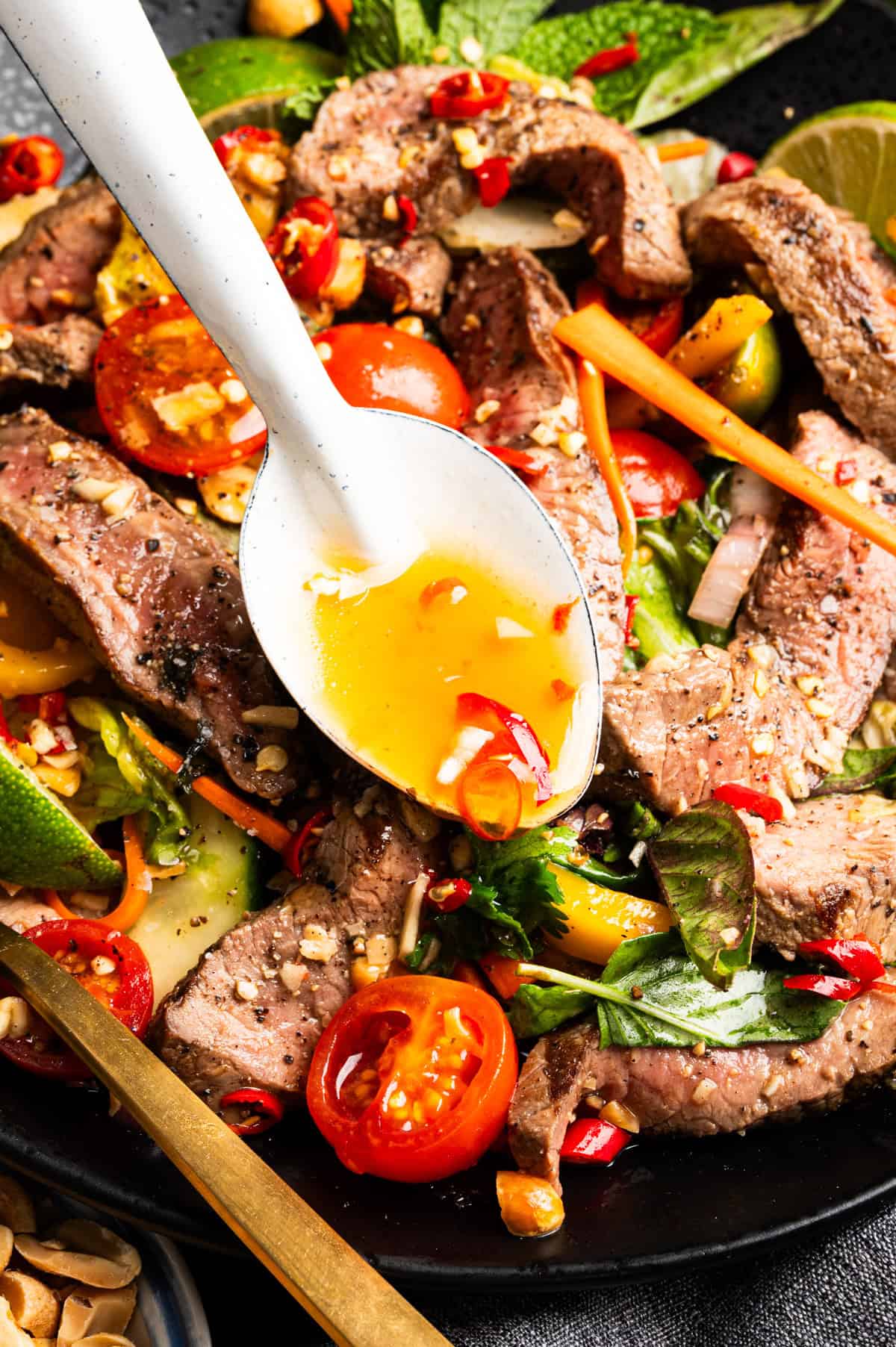 Thai Beef Salad A close-up of a vibrant steak salad with cherry tomatoes, herbs, carrot strips, and chili slices, topped with a spicy dressing being spooned over the dish.