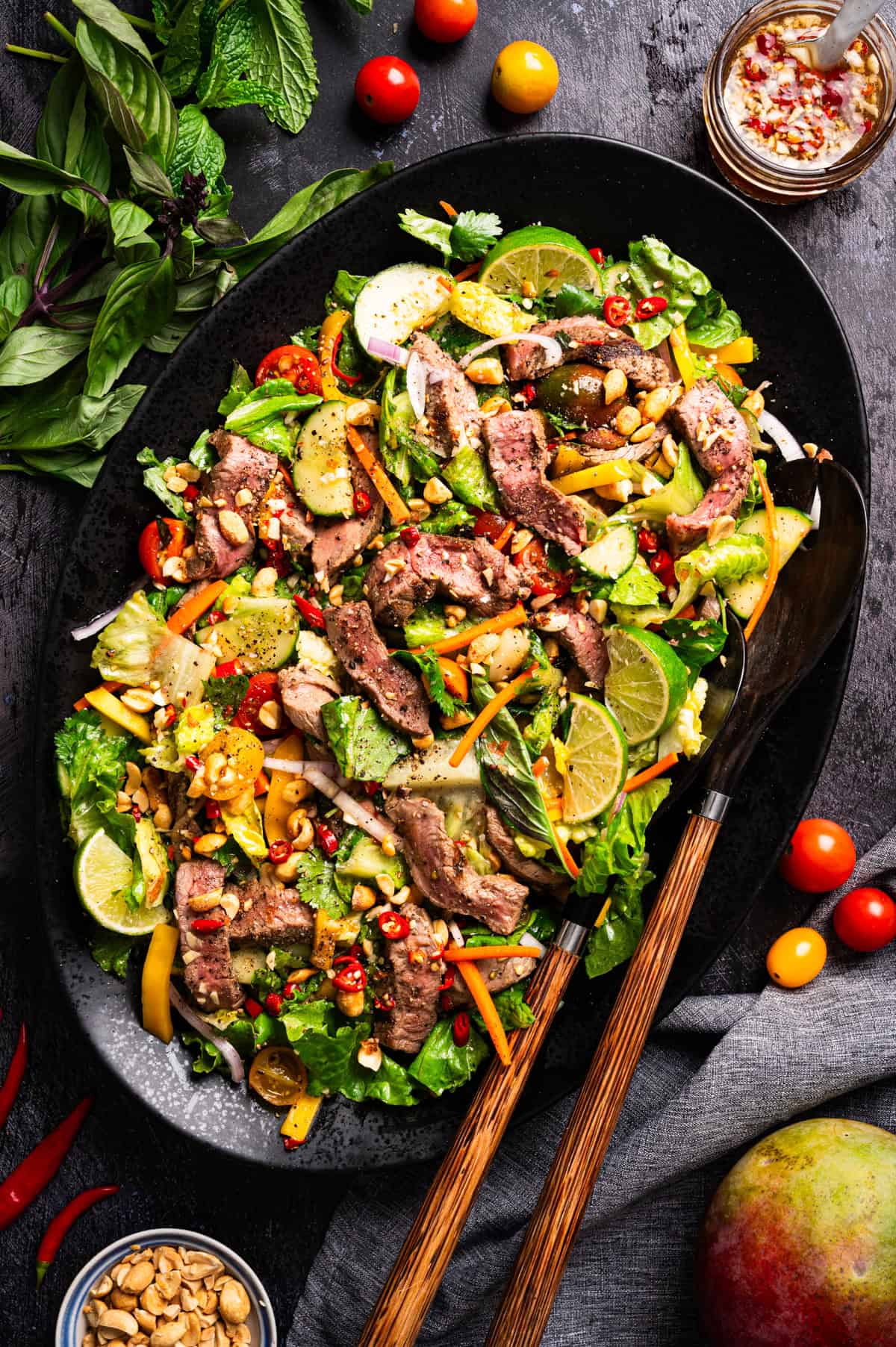 Thai Beef Salad A black platter with sliced steak on a colorful salad of greens, bell peppers, cucumbers, herbs, cherry tomatoes, peanuts, and lime wedges.