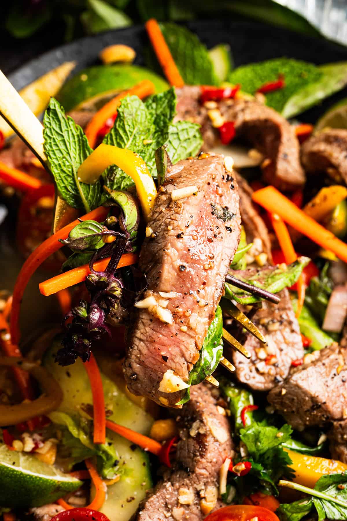 Thai Beef Salad Close-up of a fork holding a piece of seared beef above a colorful salad with carrots, greens, mint, peppers, and lime slices, garnished with herbs and spices. The meat appears juicy and tender.