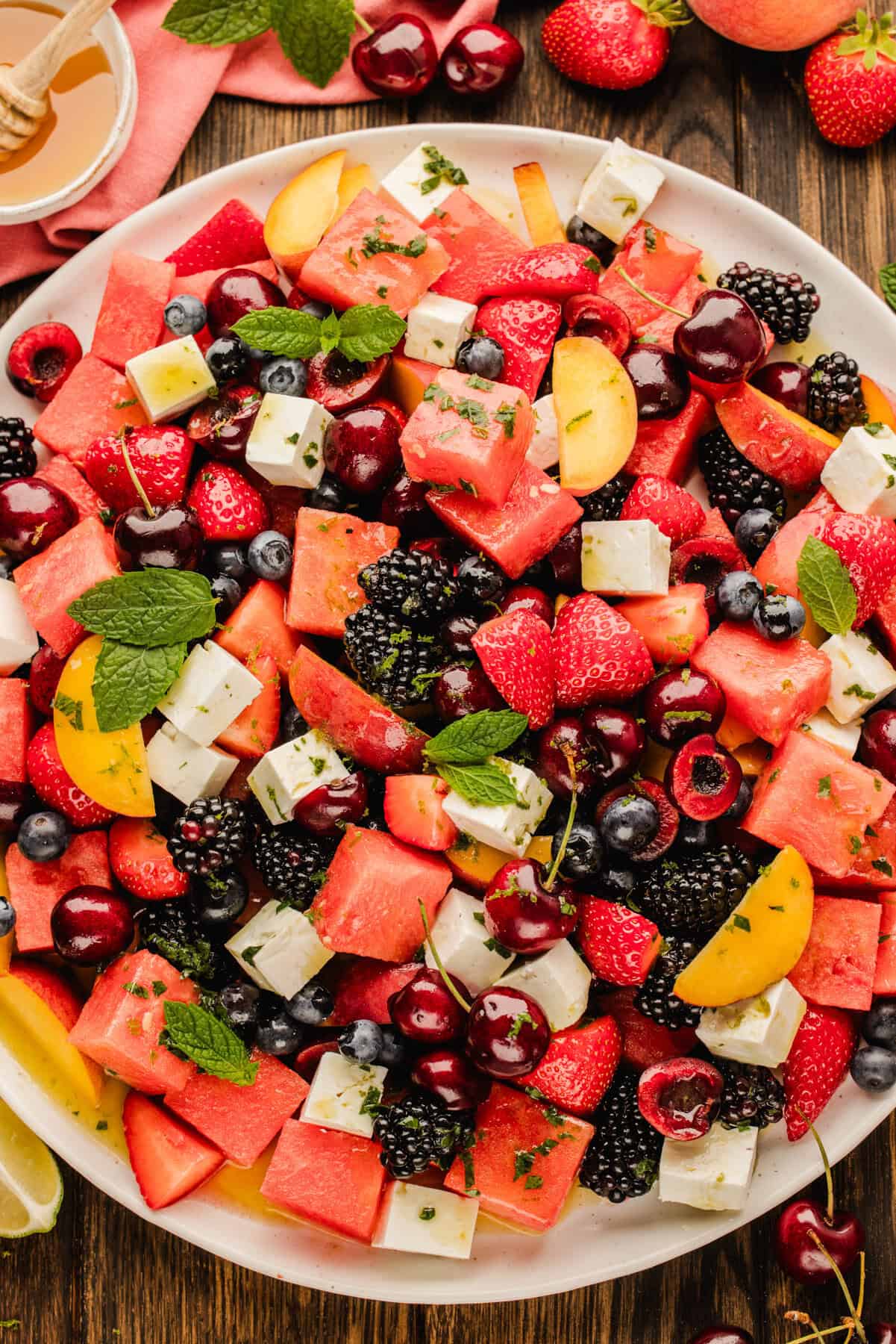 A colorful fruit salad with watermelon, strawberries, cherries, blackberries, peach slices, cubes of feta cheese, and fresh mint leaves on a white plate, surrounded by extra fruit on a wooden table.