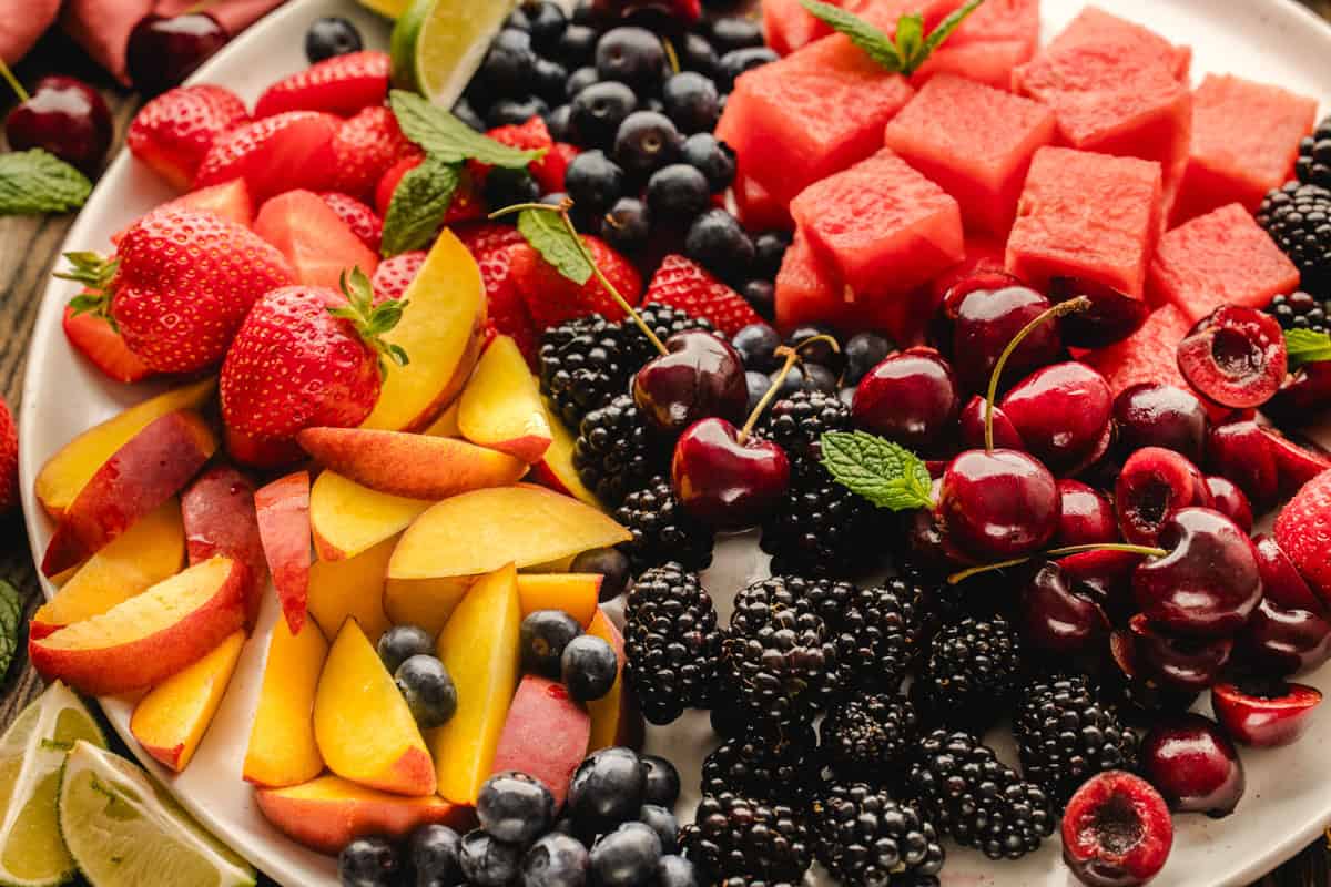 A plate filled with fresh fruits including strawberries, sliced peaches, watermelon cubes, cherries, blackberries, blueberries, and lime wedges, garnished with mint leaves.