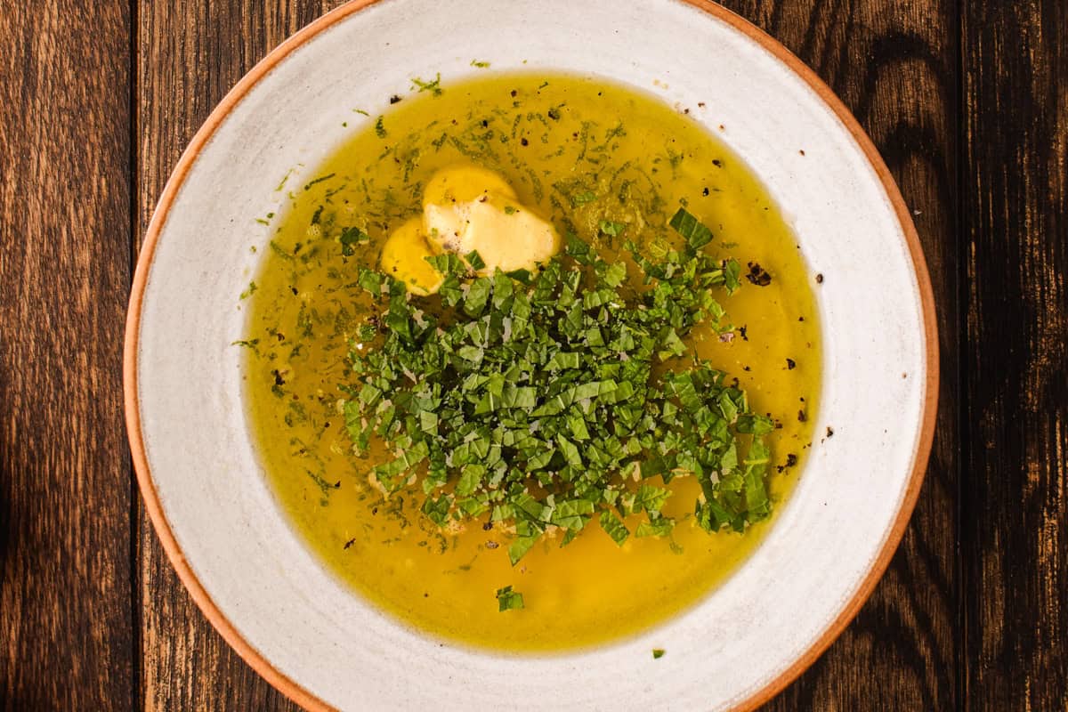 A shallow white bowl filled with olive oil, chopped fresh herbs, cracked black pepper, and a dollop of mustard, set on a wooden table.