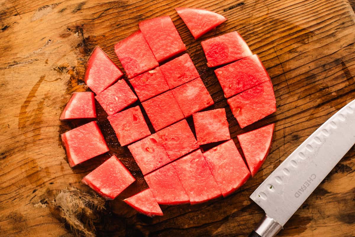 A round slice of watermelon cut into small square pieces sits on a wooden cutting board, with a stainless steel knife placed nearby.
