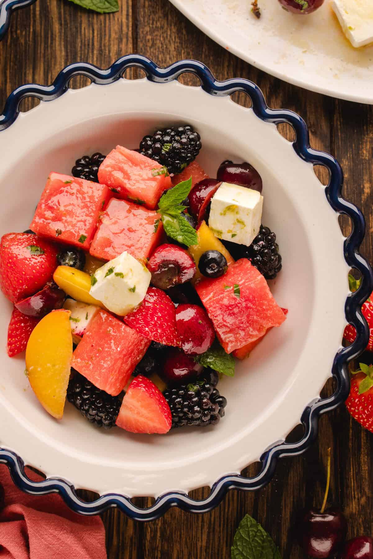 A bowl filled with a colorful fruit salad containing watermelon cubes, strawberries, blackberries, cherries, peach slices, blueberries, feta cheese cubes, and fresh mint leaves on a wooden table.