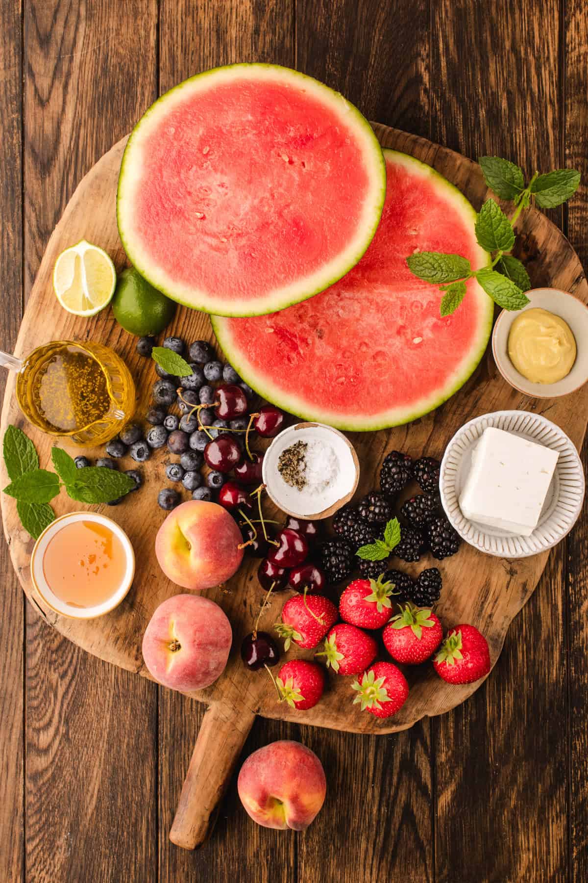 A wooden board with sliced watermelon, peaches, strawberries, blackberries, cherries, blueberries, lemon, lime, feta cheese, honey, mustard, salt, pepper, olive oil, and mint leaves, on a rustic wooden table.