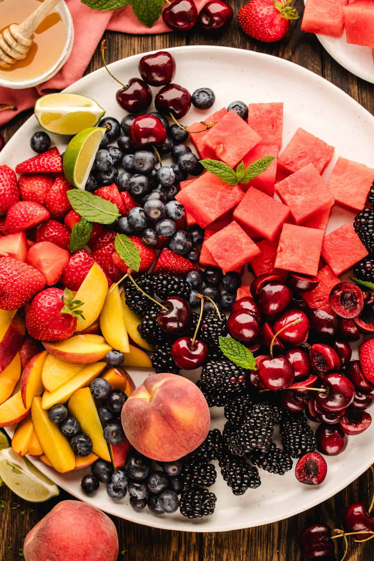 A white plate filled with fresh fruits including watermelon cubes, strawberries, cherries, blueberries, blackberries, peach slices, and lime wedges, surrounded by whole peaches and a honey dipper on a rustic wooden table.