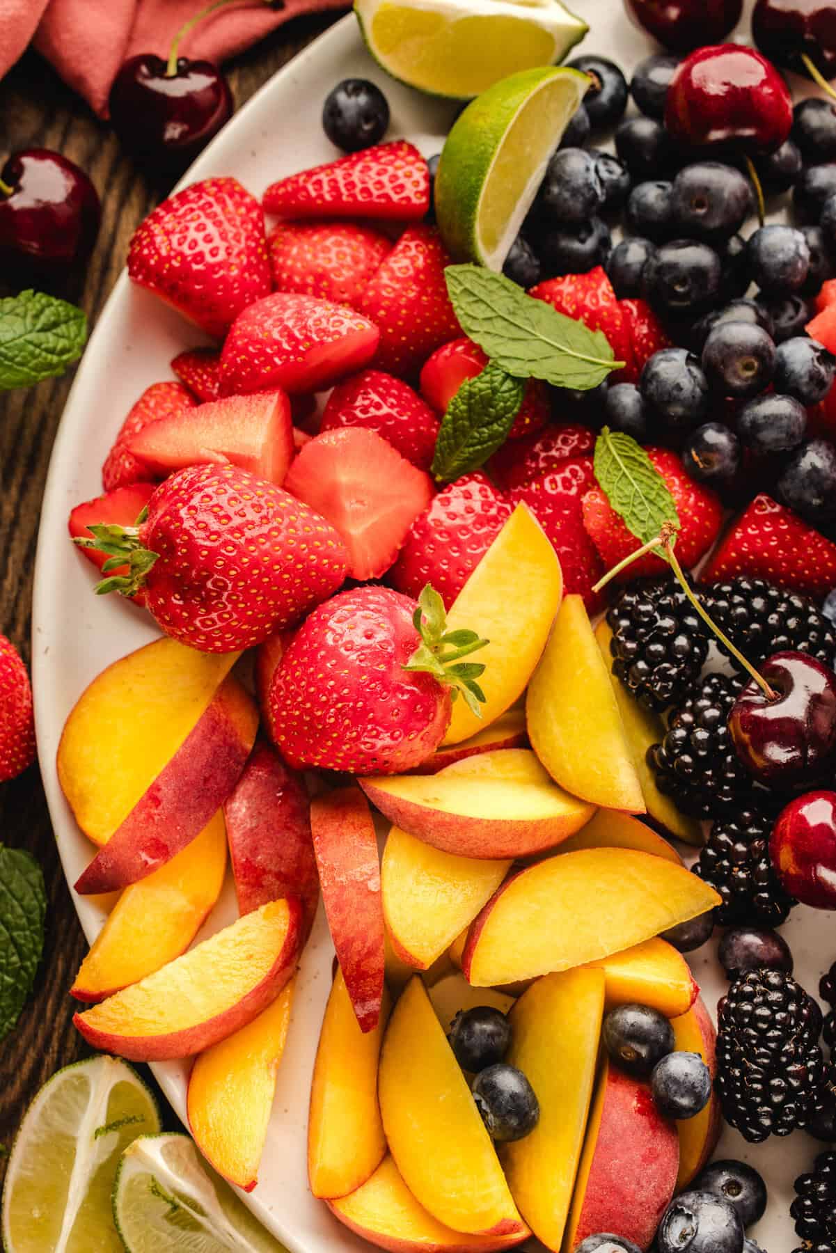 A close-up of a fruit platter featuring sliced peaches, whole strawberries, blackberries, blueberries, cherries, and lime wedges, garnished with fresh mint leaves on a white plate.