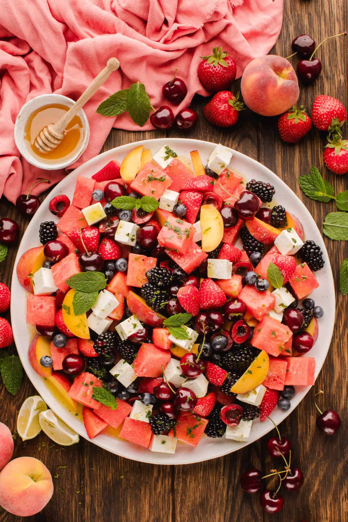 A vibrant fruit salad on a white plate with watermelon, peaches, blackberries, strawberries, cherries, feta cubes, and mint leaves. Surrounding the plate are fresh fruits, honey, and a pink cloth on a wooden table.