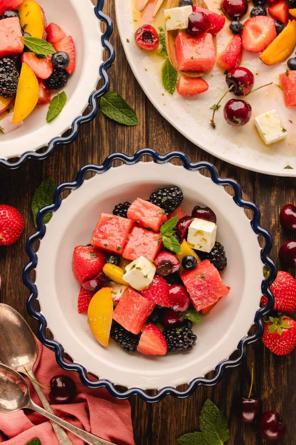 A bowl of fruit salad with watermelon, strawberries, cherries, blackberries, peach slices, mint leaves, and cubes of cheese on a wooden table beside a plate and napkin.