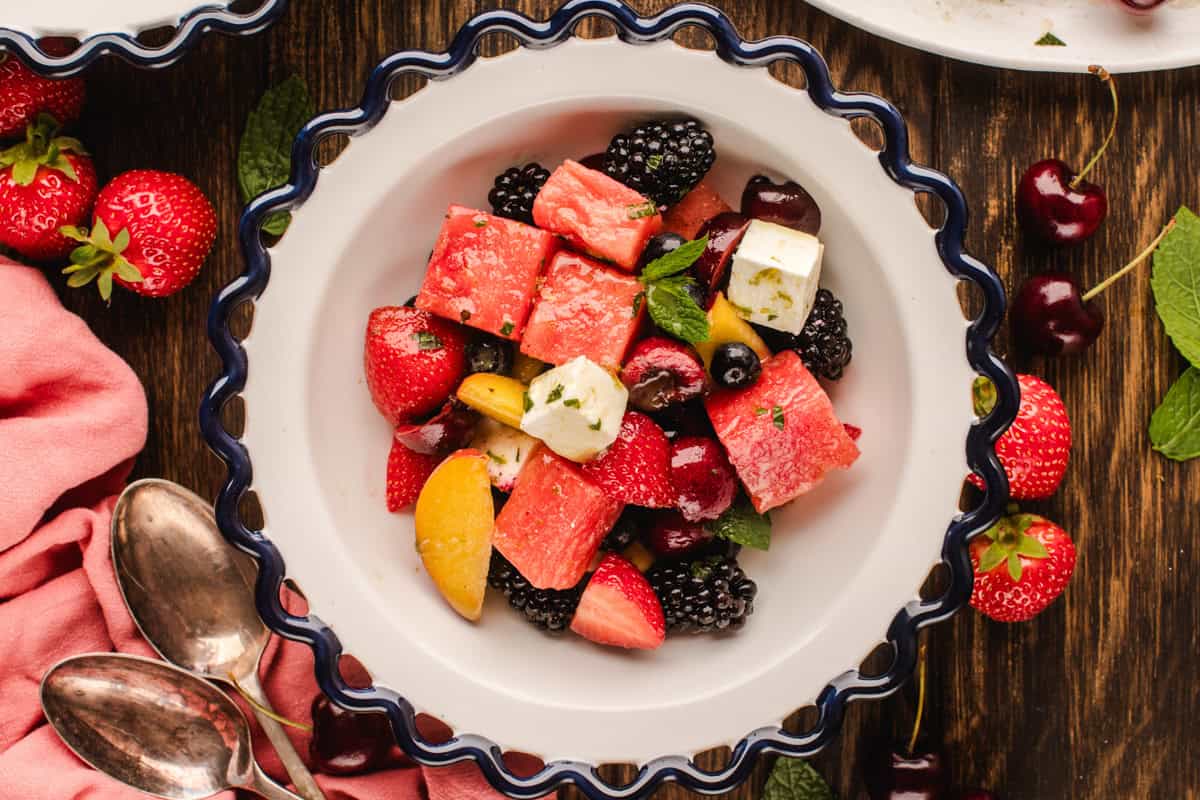 A white scalloped bowl filled with a colorful fruit salad, including watermelon, strawberries, blackberries, cherries, peaches, and feta cheese, garnished with mint leaves. The bowl is on a wooden table with scattered fruit.