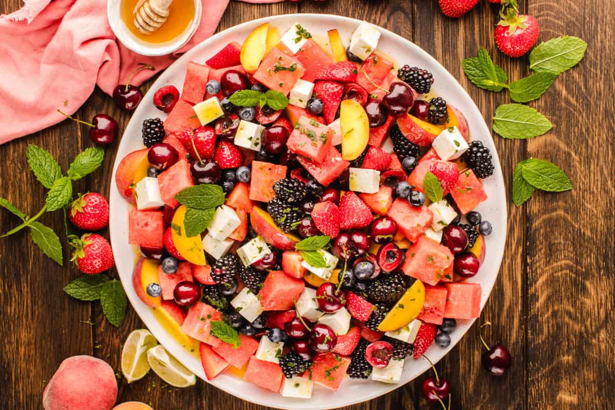 A large white plate filled with a colorful fruit salad, including watermelon, strawberries, blackberries, cherries, blueberries, peach slices, feta cheese cubes, and fresh mint leaves, on a wooden table with honey and lime nearby.
