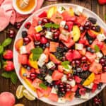 Watermelon salad on a white plate with strawberries, cherries, blackberries, blueberries, peach slices, feta cheese cubes, and mint leaves, surrounded by fresh fruit, lime wedges, and a bowl of honey.