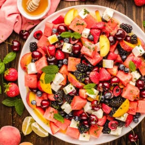 Watermelon salad on a white plate with strawberries, cherries, blackberries, blueberries, peach slices, feta cheese cubes, and mint leaves, surrounded by fresh fruit, lime wedges, and a bowl of honey.