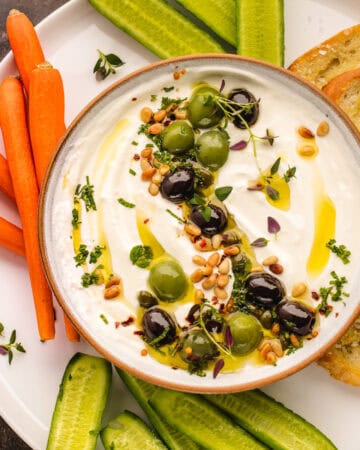 A bowl of creamy whipped feta dip topped with green and black olives, pine nuts, herbs, and olive oil, surrounded by cucumber slices, carrot sticks, and toasted bread on a white plate.