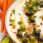 A bowl of creamy whipped feta dip garnished with green and black olives, pine nuts, chopped herbs, and a drizzle of olive oil, with carrot sticks and cucumber slices on the side.