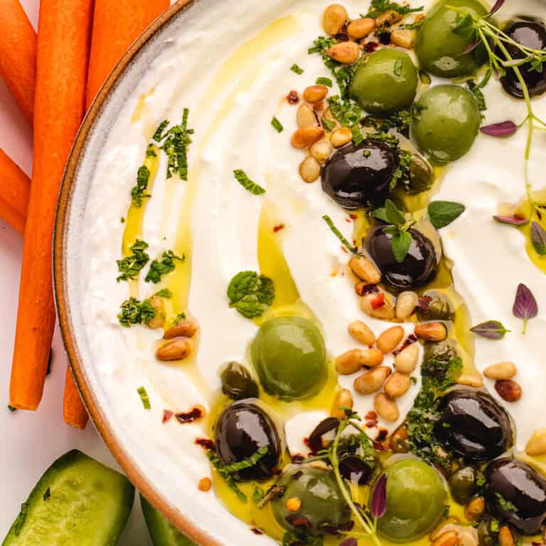 A bowl of creamy whipped feta dip garnished with green and black olives, pine nuts, chopped herbs, and a drizzle of olive oil, with carrot sticks and cucumber slices on the side.