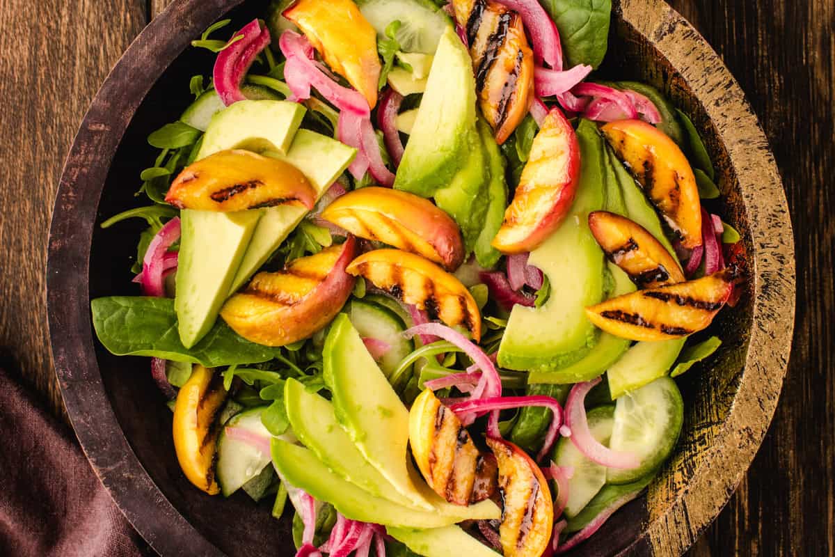 Salad Assembly A wooden bowl filled with salad containing grilled peach slices, avocado, cucumber, pickled red onions, and leafy greens on a rustic wooden table.