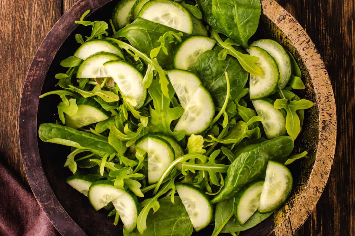 Salad Assembly A wooden bowl filled with a fresh salad of sliced cucumbers, spinach leaves, and arugula, set on a dark wooden table.
