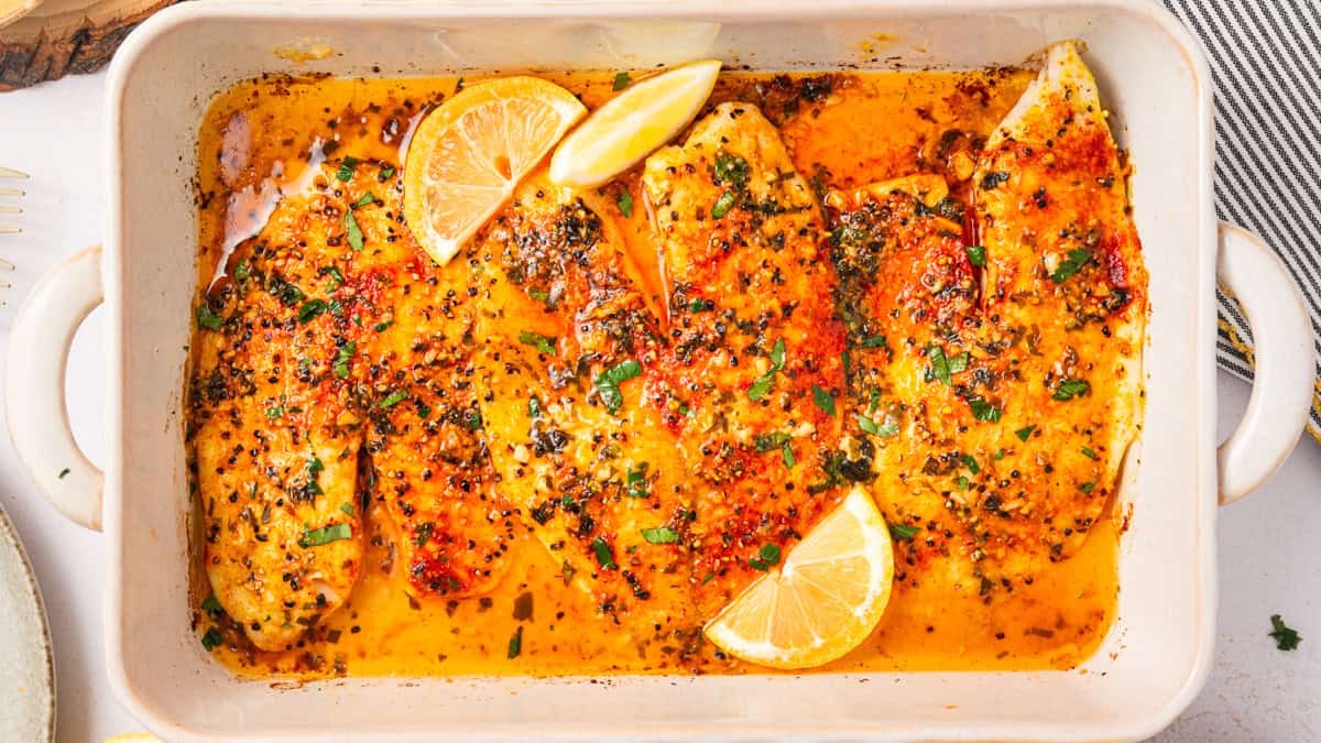 Lemon Pepper Baked Tilapia (with Lemon Butter Garlic Sauce) Baked tilapia fillets in a white dish, seasoned with herbs and spices, garnished with lemon wedges and fresh herbs, sitting in a golden, flavorful sauce.