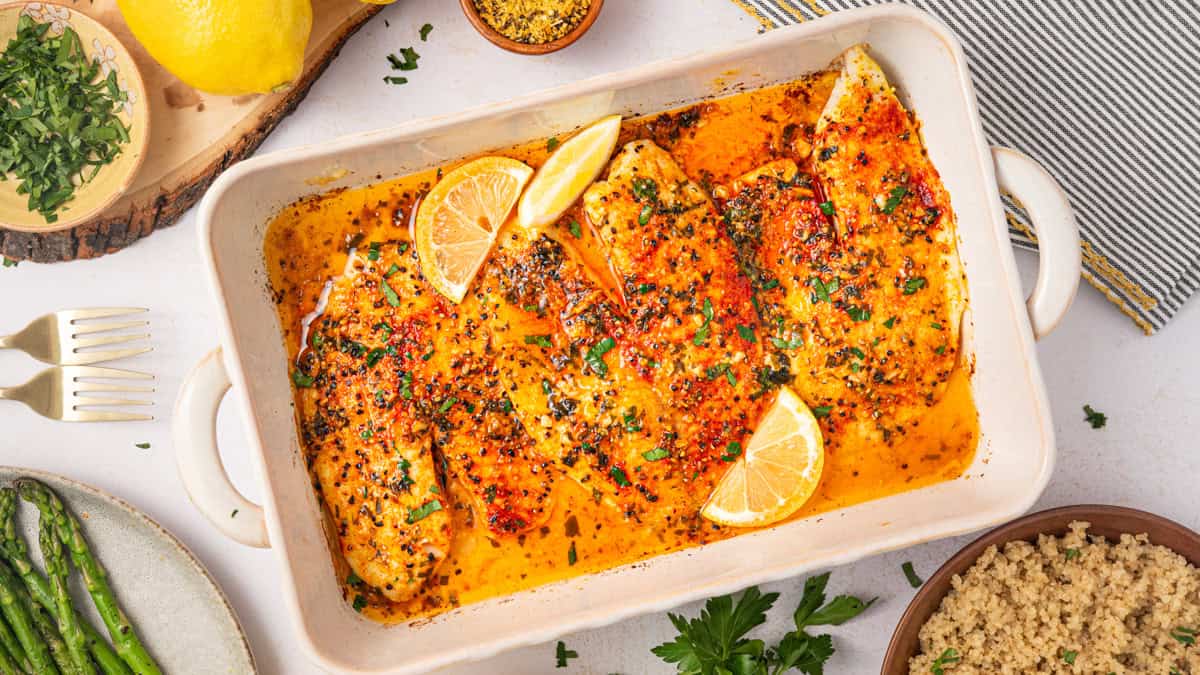 Lemon Pepper Baked Tilapia (with Lemon Butter Garlic Sauce) Baked tilapia filets in a white dish, garnished with lemon slices and chopped herbs, surrounded by bowls of parsley, quinoa, asparagus, and a halved lemon on a white surface.