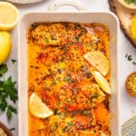 Baked tilapia fillets in a creamy, seasoned sauce, garnished with lemon wedges and fresh herbs, in a white baking dish.