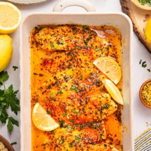 Baked tilapia fillets in a creamy, seasoned sauce, garnished with lemon wedges and fresh herbs, in a white baking dish.