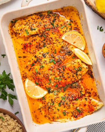 Baked tilapia fillets in a white dish, topped with herbs and spices, garnished with lemon wedges.