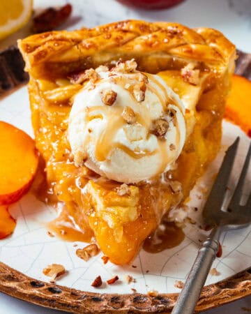 A slice of Peach Pie topped with a scoop of vanilla ice cream, caramel sauce, and chopped nuts, served on a plate with peach slices and a fork beside it.