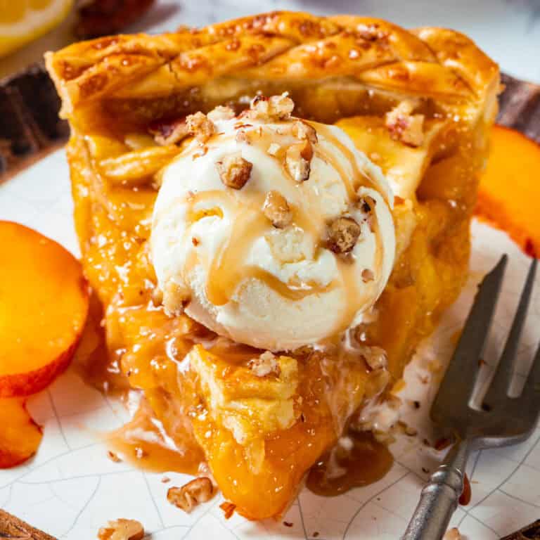 A slice of Peach Pie topped with a scoop of vanilla ice cream, caramel drizzle, and chopped pecans, served on a plate with fresh peach slices and a fork.