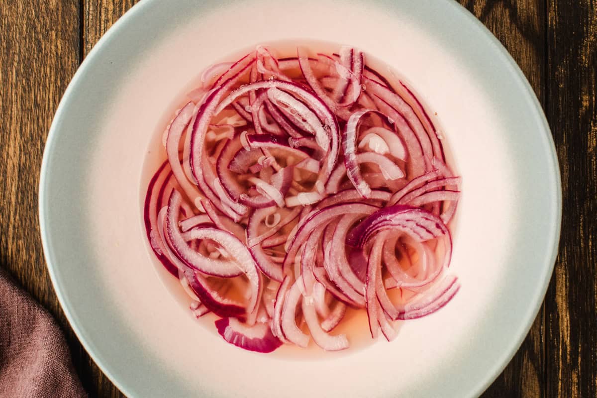 Pickled Onions for Blackberry Peach Salad Thinly sliced red onions soaking in a light pink liquid, likely vinegar, in a white shallow bowl on a wooden table.