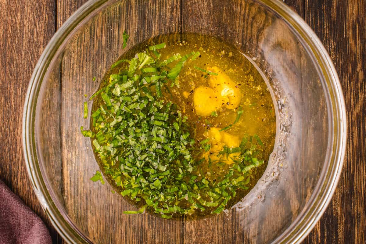 Salad Dressing for Blackberry Peach Salad A glass bowl on a wooden surface contains olive oil, chopped fresh herbs, minced garlic, and spices, ready to be mixed for a marinade or dressing.