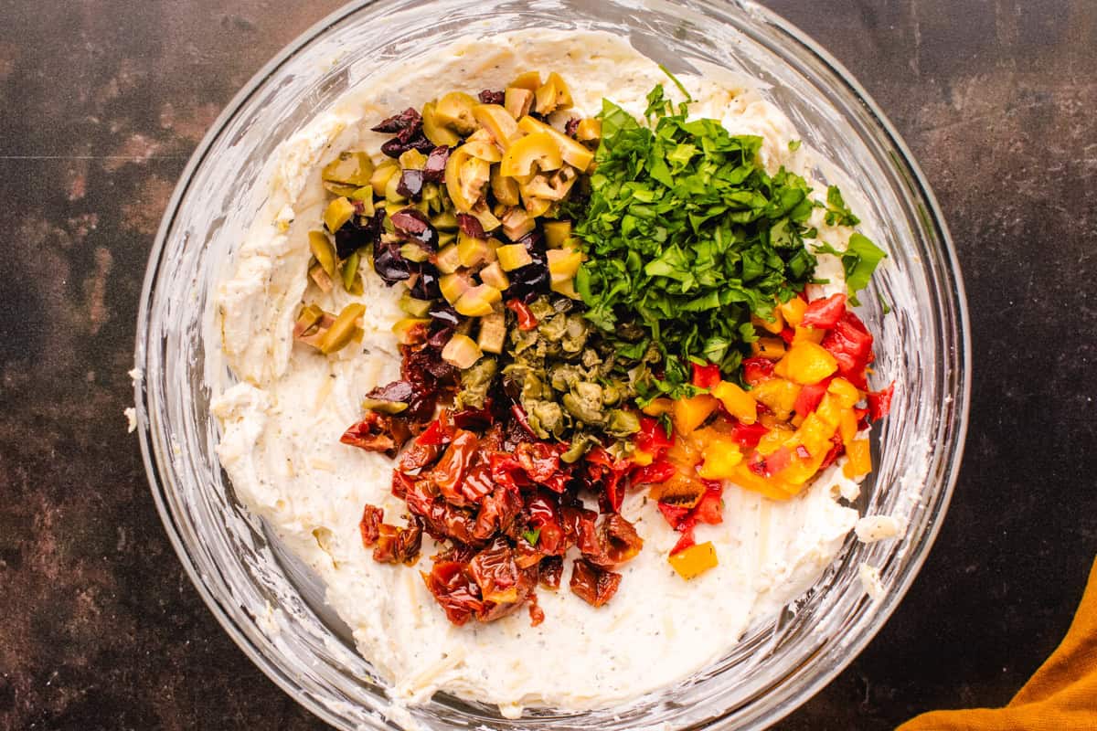 A glass bowl with a creamy base topped with chopped olives, parsley, capers, sun-dried tomatoes, and diced bell peppers, ready to be mixed together.