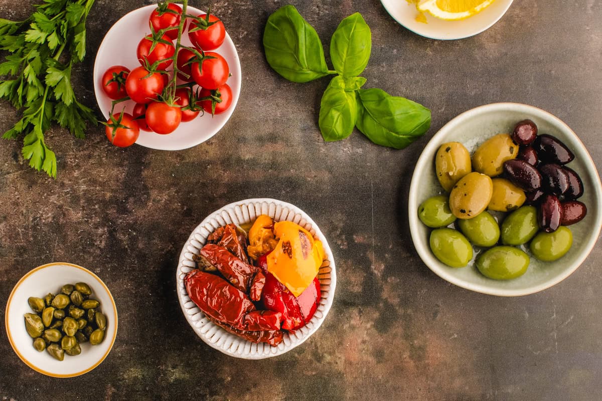 A flat lay of fresh basil, cherry tomatoes, olives, sun-dried tomatoes, capers, and lemon slices arranged in bowls on a rustic surface.