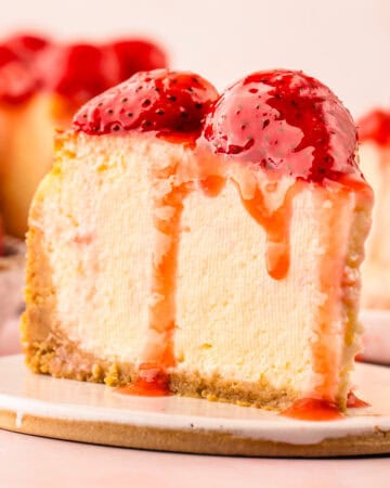 A slice of creamy cheesecake topped with glossy strawberries and strawberry sauce sits on a white plate, with more cheesecake in the background.