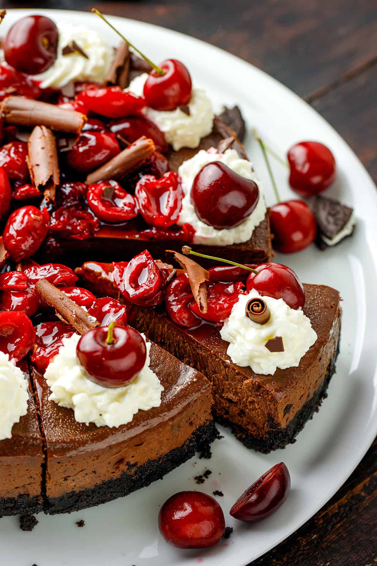 Black Forest Cheesecake A chocolate cheesecake topped with whipped cream, fresh cherries, chocolate curls, and cherry sauce, served on a white plate with whole cherries around it.