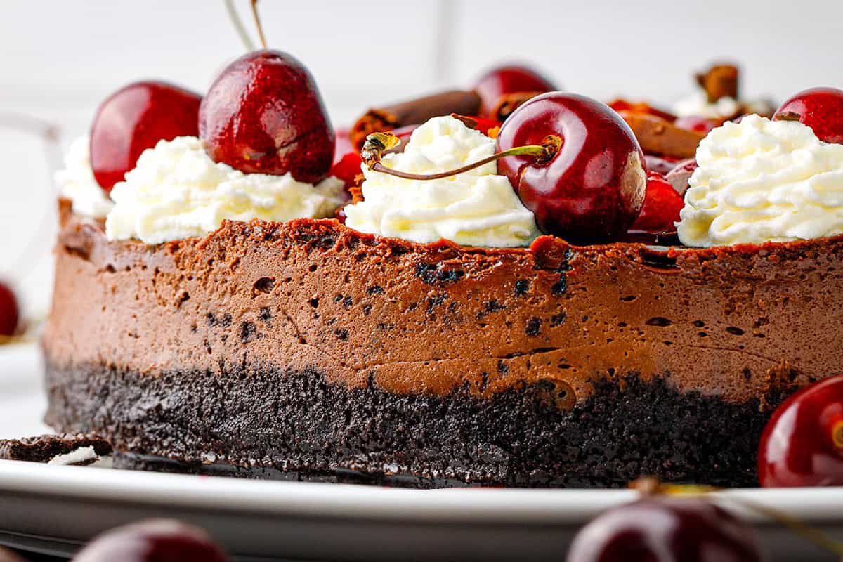 Black Forest Cheesecake A close-up of a chocolate cheesecake with a dark crust, topped with whipped cream and fresh cherries on a white plate.
