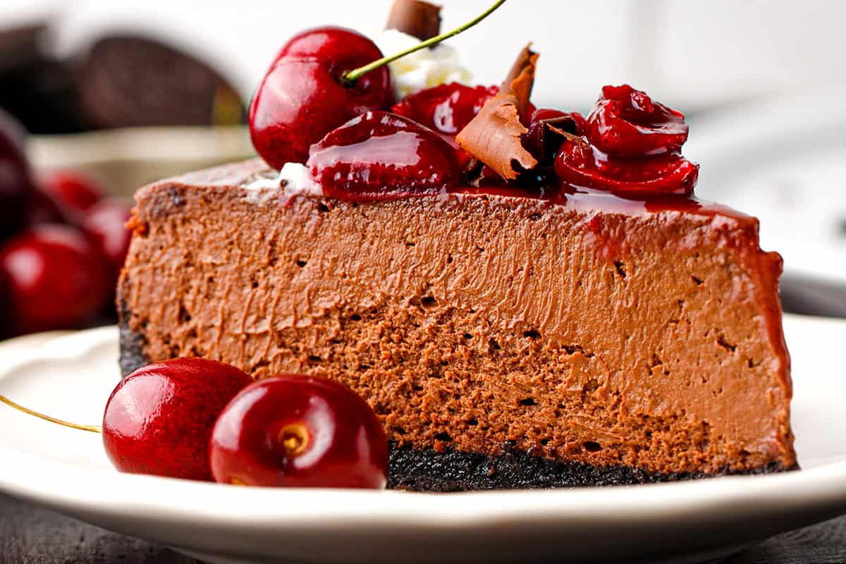 Black Forest Cheesecake A slice of chocolate cheesecake on a white plate, topped with glossy cherries and chocolate shavings, with a chocolate cookie crust and more cherries around the plate.